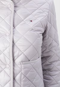 Light grey quilted jacket with diamond pattern, featuring a button closure and a small logo. It has a side pocket with a matching trim.