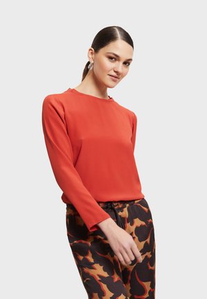Rode blouse met lange mouwen van glad materiaal, gecombineerd met gemusterde zwarte en bruine broek met een trekkoord in de taille.