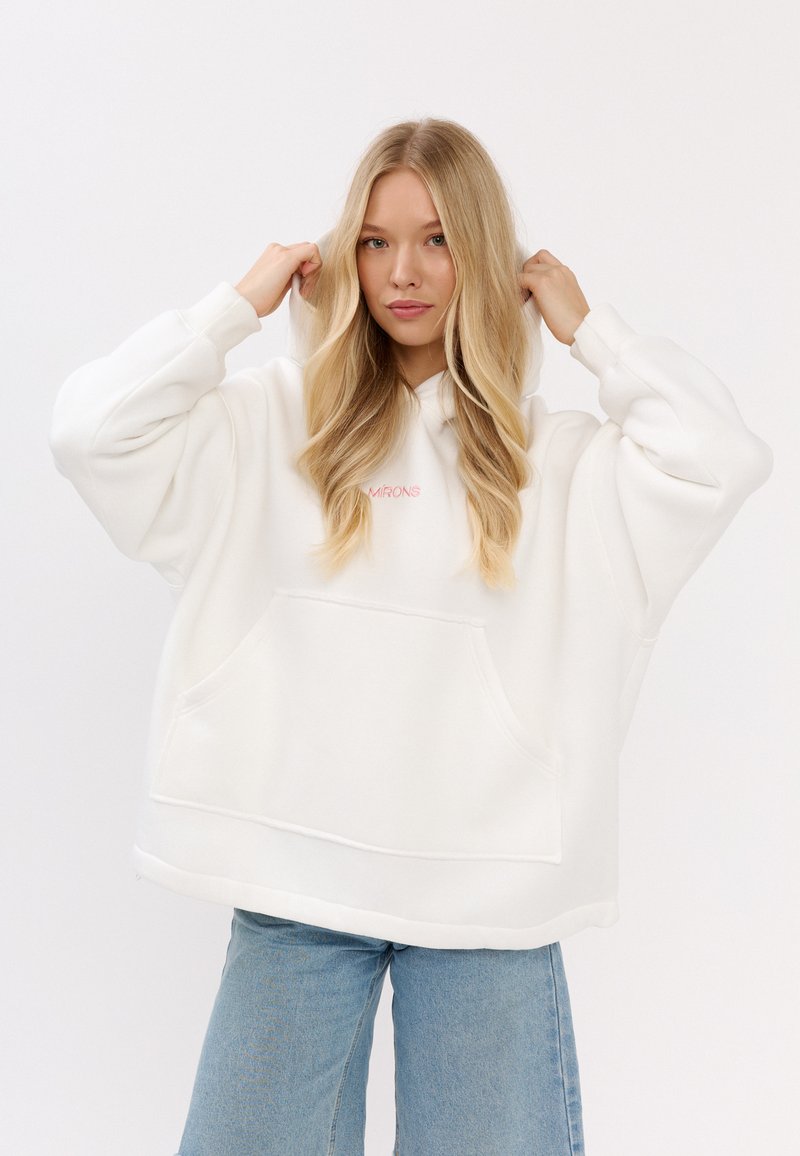 Witte oversized hoodie van zachte stof met een buidelzak en een roze logo. Model draagt lichtblauwe jeans, met de armen omhoog bij de capuchon.