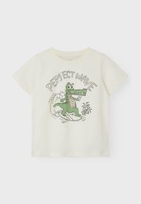 Cream short-sleeve t-shirt featuring a green crocodile surfing with the text "PERFECT WAVE" in black above, with small palm trees in the design.
