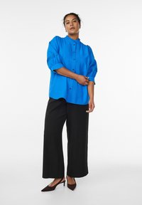 Sinine nööpidega särgi, millel on punnitatud varrukad ja peened voltimisdetailid. Sobitatud mustade laiade pükstega ja pruunide teravate kingadega.