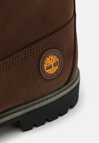 Bruine suède laars met een rond bruine logo patch met een oranje boomontwerp. De zool is zwart met een robuuste textuur voor grip.