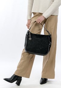 Persona, kas valkā bēša krāsas bikses un melnas augstpapēžu zābakus, tur melnu ādas somu ar sudraba piekariņu pie siksniņas.