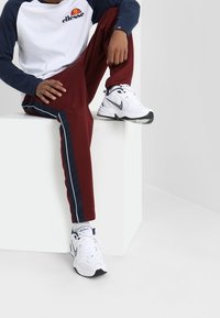 Bílá a modrá košile s dlouhým rukávem s logem "ellesse", červené tepláky s námořnickými bočními pruhy a bílé tenisky Nike s černými akcenty.