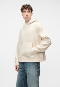 Νεαρός άνδρας με σγουρά μαλλιά, φορώντας εκρού φαρδιά φούτερ με κουκούλα και μπλε τζιν παντελόνι με φαρδιά πόδια, στέκεται μπροστά σε απλό φωτεινό φόντο.