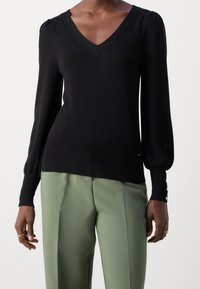 Suéter negro de cuello en V con mangas largas y abullonadas, y puños acanalados. Presenta acentos de botones dorados en los puños. Combinado con pantalones verde claro.