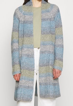 Cárdigan largo de punto con rayas horizontales en azul, gris, verde y blanco, llevado sobre una camiseta verde y pantalones blancos.