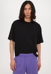 Camiseta negra de gran tamaño hecha de tela suave, con hombros caídos y un detalle de etiqueta en el dobladillo, combinada con pantalones cortos de cintura elástica morados.