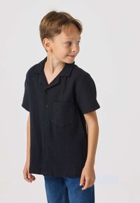 Sort kortærmet skjorte med knapper og en lomme, lavet af tekstureret stof; kombineret med blå denimjeans.