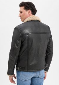Schwarze Lederjacke mit einem beigen Schaffellkragen, ausgestattet mit einer glatten Textur und seitlichen Schnallenakzenten. Von hinten gezeigt.