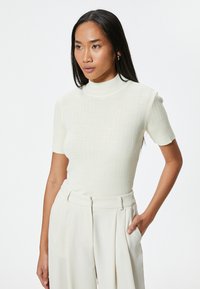 Top corto de punto acanalado color crema con cuello alto. La tela parece suave y tiene una silueta ajustada. Combinado con pantalones de color claro.