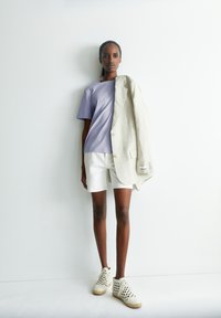 Hellblaues T-Shirt mit kurzen Ärmeln, weiße Shorts, beiger Leinenblazer über die Schulter gelegt und beiger Schuh mit Ausschnitten und gewebter Textur.