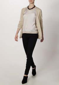 Beige cardigan over a light silk blouse, paired with black slim-fit pants and ankle booties, featuring a bold statement necklace.