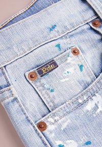 Džínové lacláče s světle modrým praním, s detaily rozstříkané barvy. Hnědé kovové knoflíky a značková nášivka s "Polo" v žluté.