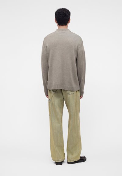 Homme portant un pull en maille taupe et un pantalon ample vert clair, dos tourné contre un fond blanc uni.