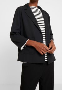 Blazer noir avec un col cranté, une poche avant et une fermeture à un bouton, superposé à une chemise rayée avec un bord blanc.