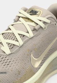 Μπεζ αθλητικό παπούτσι με υφή στην επιφάνεια, ανοιχτόκίτρινα κορδόνια και μεταλλικό χρυσό λογότυπο swoosh. Διαθέτει διακριτικές λεπτομέρειες με μικρές βούλες στην επιφάνεια.