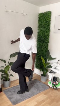Schwarze Hosen und ein weißes T-Shirt, getragen von einer Person, die ihre Hose anpasst; schwarze Schuhe, grauer strukturierter Teppich, grüne Pflanzen im Hintergrund.