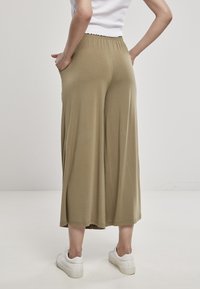 Person som står med händerna i fickorna, iförd beige vida byxor, vita sneakers och en vit ärmlös topp, vänd bort från kameran mot en neutral bakgrund.