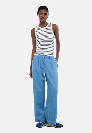 Femme portant un haut blanc sans manches à rayures noires, un jean pattes d'éléphant bleu clair et des baskets bleues, debout avec une main dans sa poche.