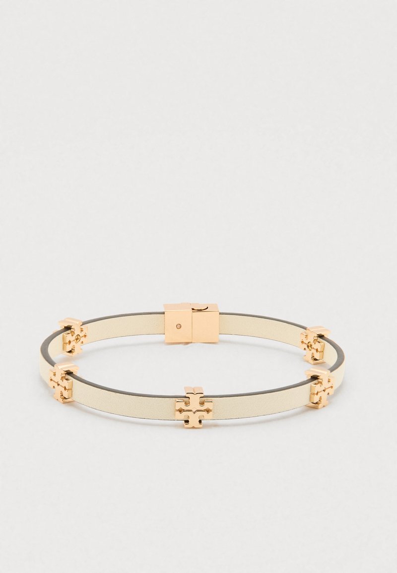 Μπεζ δερμάτινο βραχιόλι με χρυσές σταυροειδείς λεπτομέρειες ομοιόμορφα τοποθετημένες και ορθογώνιο χρυσό κούμπωμα σε απλό φόντο.