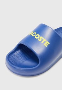 Blue plastic slide sandal with open toe design, featuring a textured insole and the word "LACOSTE" in yellow on the upper strap.