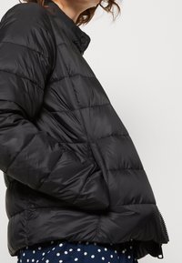 Giacca a vento nera, design trapuntato con colletto alto, dotata di chiusura a zip e tasche laterali. Texture liscia e lucida.