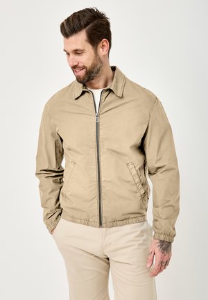 Man with beard wearing beige zip-up jacket and light-colored pants, looking down with hands in pockets, showing wrist tattoos.