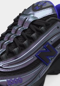 Gros plan sur une chaussure de sport New Balance 1000 noire et irisée avec des accents violets et un laçage détaillé sur une tige texturée.