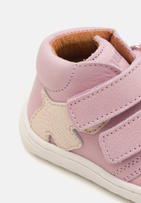 Rosa lærskode med en teksturert beige stjerneakcent, med to borrelåsremmer og en hvit gummisåle. Nærbilde av ankelområdet.