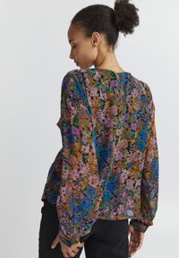 La blusa floreale presenta una stampa colorata con tonalità di blu, rosa e verde. Maniche lunghe e ampie e un tessuto leggero, leggermente trasparente.