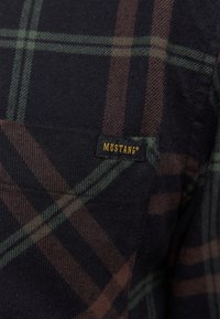 Chemise en flanelle à carreaux présentant des motifs bleu foncé, vert et marron. Comprend une poche avec une étiquette noire "MUSTANG". Tissu doux et texturé.
