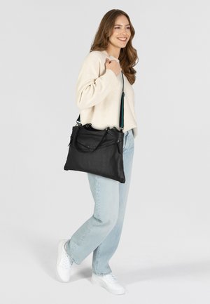 Sac à main en cuir noir avec deux poignées et une bandoulière amovible, présentant une surface texturée et une poche zippée. Porté avec un jean bleu clair.