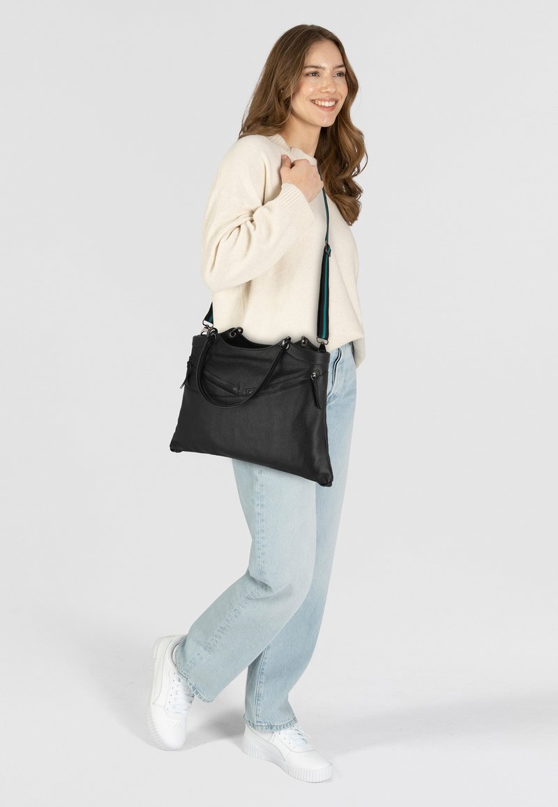 Sac à main en cuir noir avec deux poignées et une bandoulière amovible, présentant une surface texturée et une poche zippée. Porté avec un jean bleu clair.