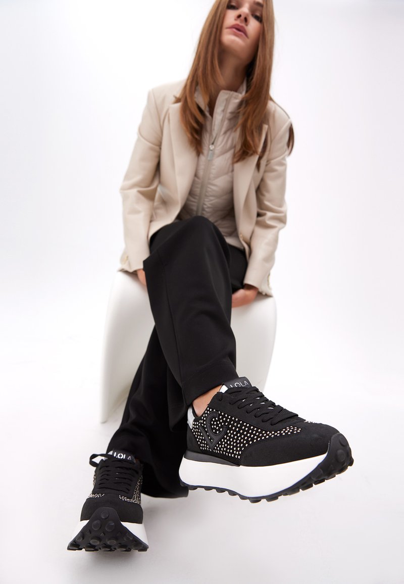 Mustat urheilukengät, joissa on teksturoitu mokkanahka päällinen, niittikoristeet, valkoinen välipohja ja kumipohja, yhdistettynä mustiin housuihin ja beigeen takkiin.