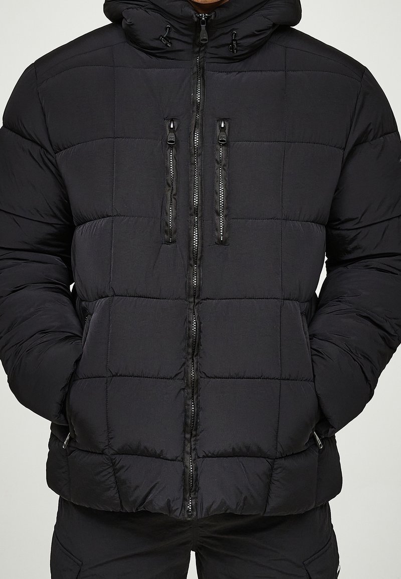 Homme portant une veste matelassée noire avec capuche, poches poitrine et latérales zippées, les mains dans les poches latérales.
