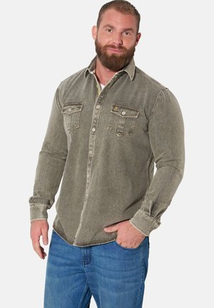 Bärtiger Mann, der ein olivgrünes Hemd mit Brusttaschen und blaue Jeans trägt, stehend mit einer Hand in der Tasche.