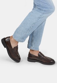 Mocassins en cuir marron avec un bout défini, des détails de couture et une semelle noire à profil bas, associés à un jean bleu clair retroussé.