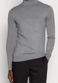 Homme portant un pull en col roulé gris et un pantalon noir, avec une main dans la poche, sur un fond uni.