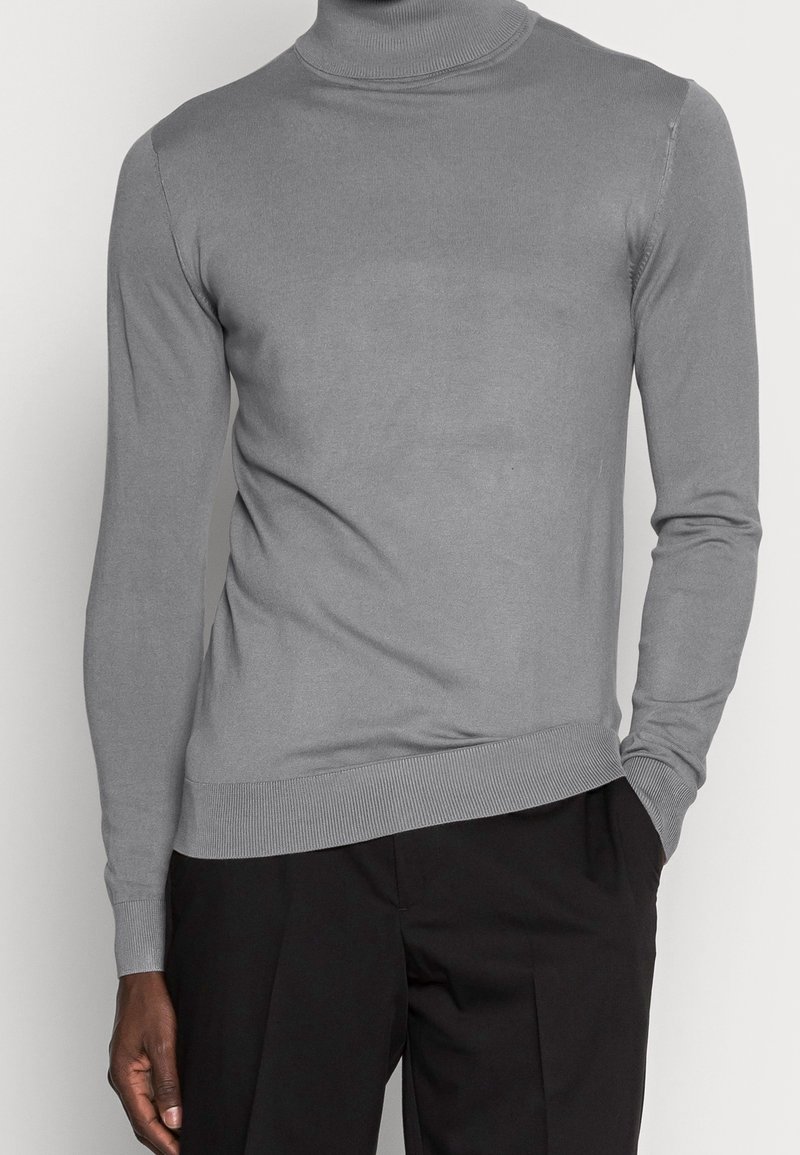 Homme portant un pull en col roulé gris et un pantalon noir, avec une main dans la poche, sur un fond uni.