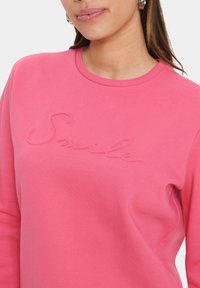 Felpa rosa con collo a girocollo e scritta "Smile" ricamata dello stesso colore. Tessuto morbido, maniche lunghe e vestibilità comoda.