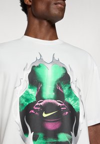 Hvid T-shirt med et centralt grafik, der viser en basketball, grønne og pink accenter samt sølvfarvede flamme-lignende detaljer.