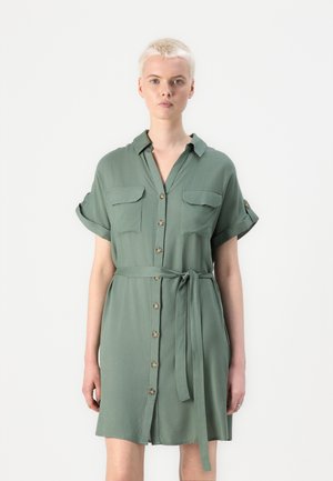 Abito camicia verde realizzato in tessuto morbido. Presenta una chiusura con bottoni frontalmente, due tasche sul petto, maniche arrotolate e una cintura da annodare.