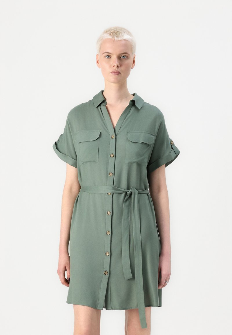 Abito camicia verde realizzato in tessuto morbido. Presenta una chiusura con bottoni frontalmente, due tasche sul petto, maniche arrotolate e una cintura da annodare.