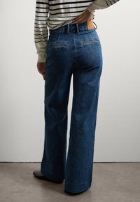 Pantalones de mezclilla de pierna ancha en azul oscuro, confeccionados con tela texturizada. Cuentan con bolsillos traseros cosidos y una etiqueta de cuero en la cintura.