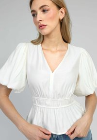 Femme portant un haut blanc à manches courtes bouffantes et taille froncée, posant devant un fond gris uni.