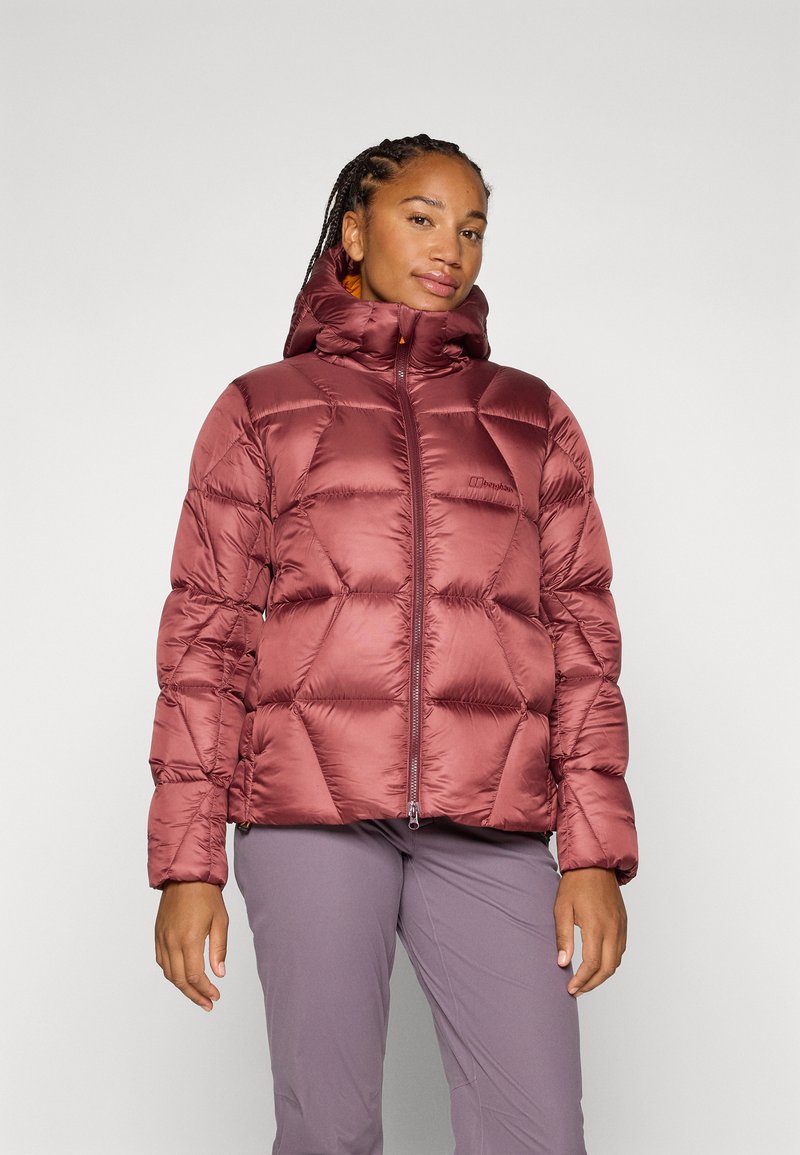 Una mujer con trenzas lleva una chaqueta de plumas burdeos y pantalones grises, de pie frente a un fondo liso con una expresión de confianza.