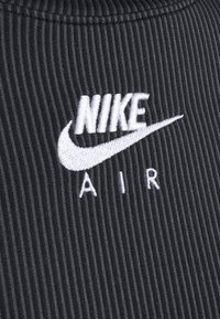 Svart ribbat tyg med en vit broderad "NIKE"-logotyp och "AIR" nedanför. Texturen är uttalad och ger djup åt designen.