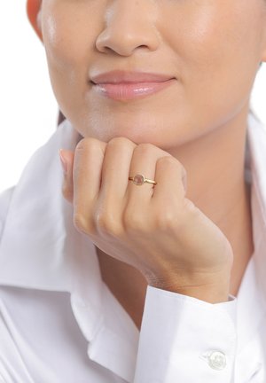 Kvinde der hviler hagen på sin knytnæve, bærer en enkel guldring med en rund sten, iført en hvid skjorte med knapper.