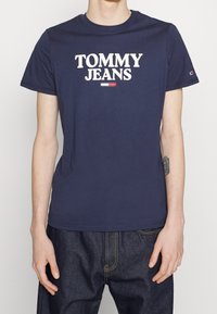 Námořnicky modré bavlněné tričko s nápisem "TOMMY JEANS" v tučných bílých písmenách. Kulatý výstřih a krátké rukávy. Klasický střih.